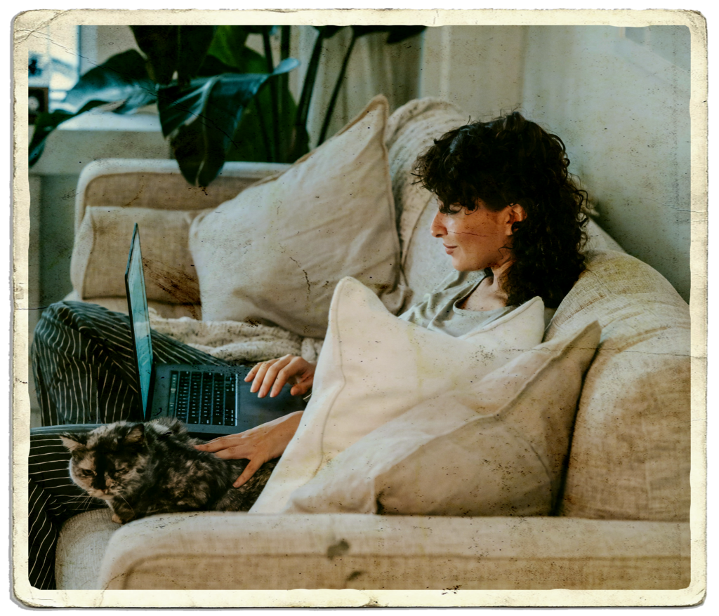 A young woman sitting on a sofa scrolling on her laptop while stroking a cat.