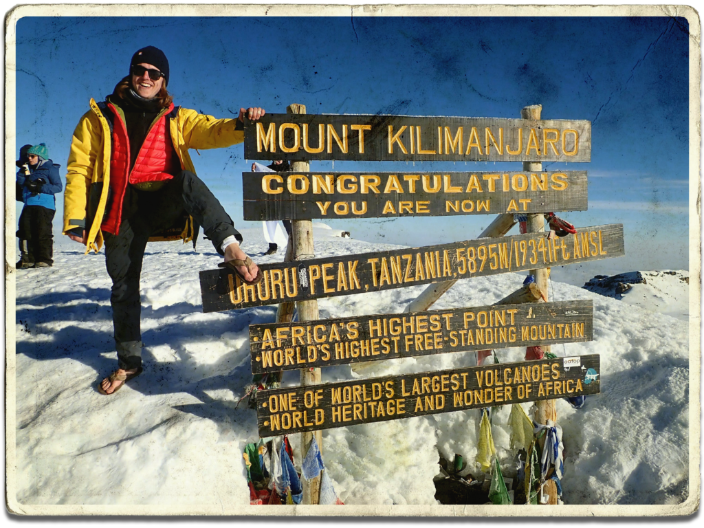 Next Stop Adventure's Content Strategist Sam Oliver posing next to the Mount Kilimanjaro summit sign wearing hiking gear and flip-flops.