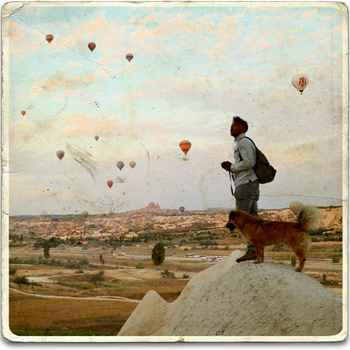 A square old school postcard with a young male adventurer and a dog stood on a boulder and watching the hot air balloons fly over Cappadocia, Turkey.