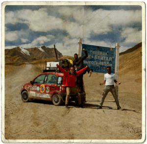 The Mongol Rally 2019 team Bristangolia, including social media travel agency Next Stop Adventures founder and creative strategist, celebrating reaching the highest point of the Pamir highway in front of a road sign and next to their Fiat Panda.